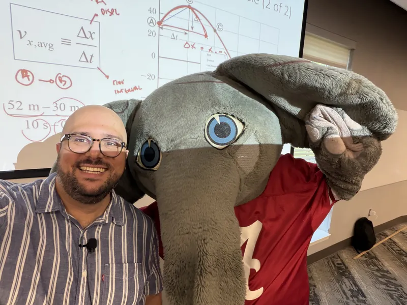 Selfie of Emanuele Usai with the Big Al elephant mascot in front of a lecture slide defining average velocity as the change in position divided by the change in time.