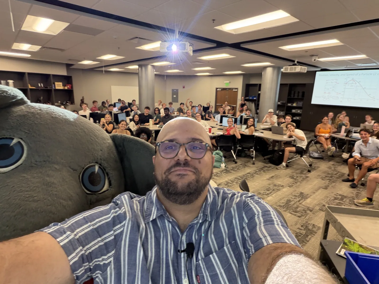 Selfie of Emanuele Usai in front of a large introductory physics class seated at group tables, with the Big Al elephant mascot beside him and a lecture slide on the screen.