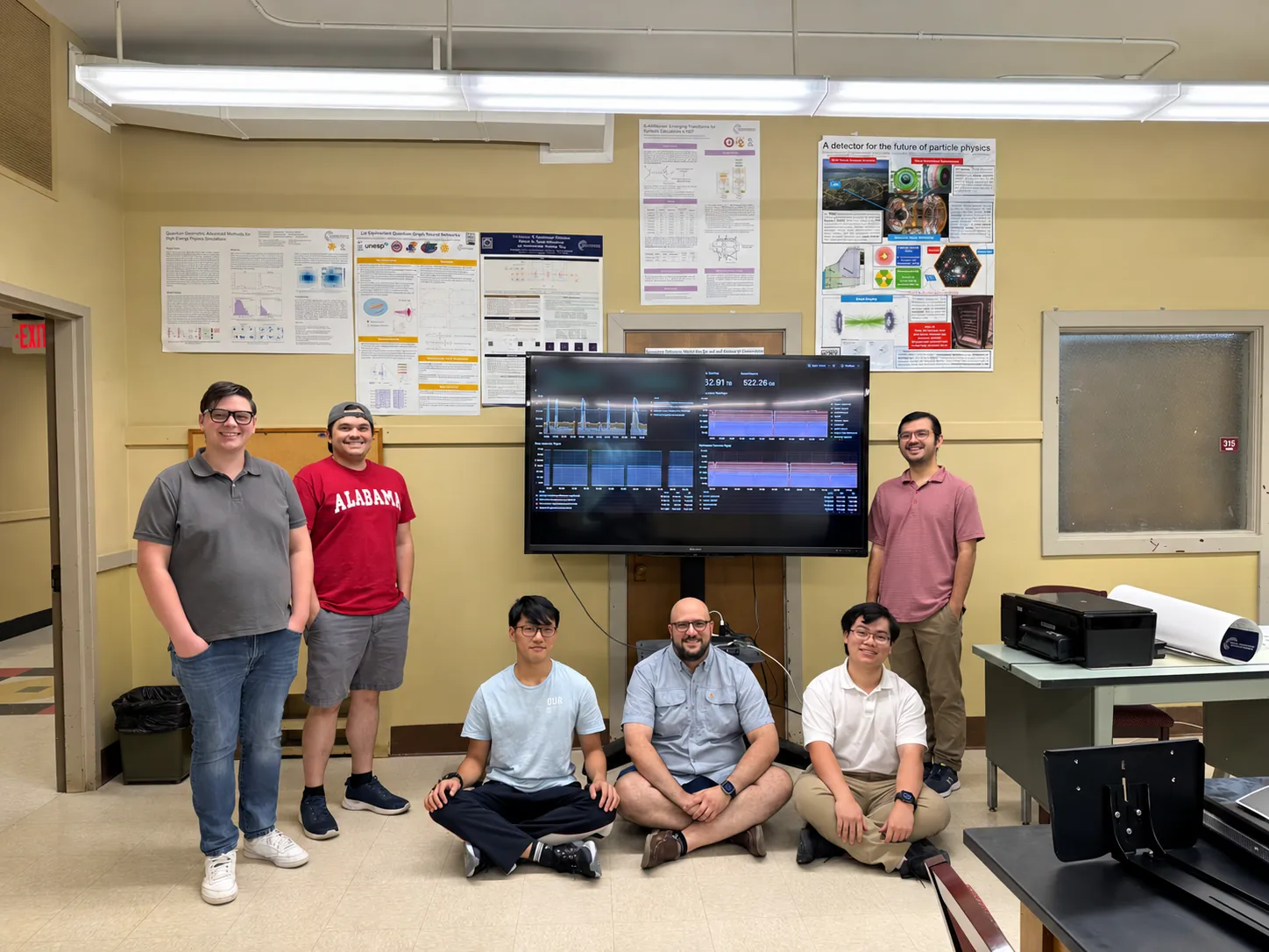 Six members of the group in the lab, three standing and three sitting on the floor, around a large screen on a stand, with research posters on the wall behind them.