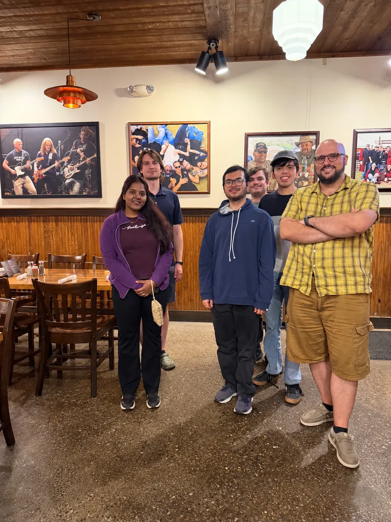 Seven people standing together in a restaurant dining room, in front of framed photographs on a wood-paneled wall.