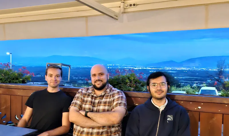 Emanuele Usai, center, sitting with two colleagues on a terrace at dusk, with a valley and city lights in the distance.
