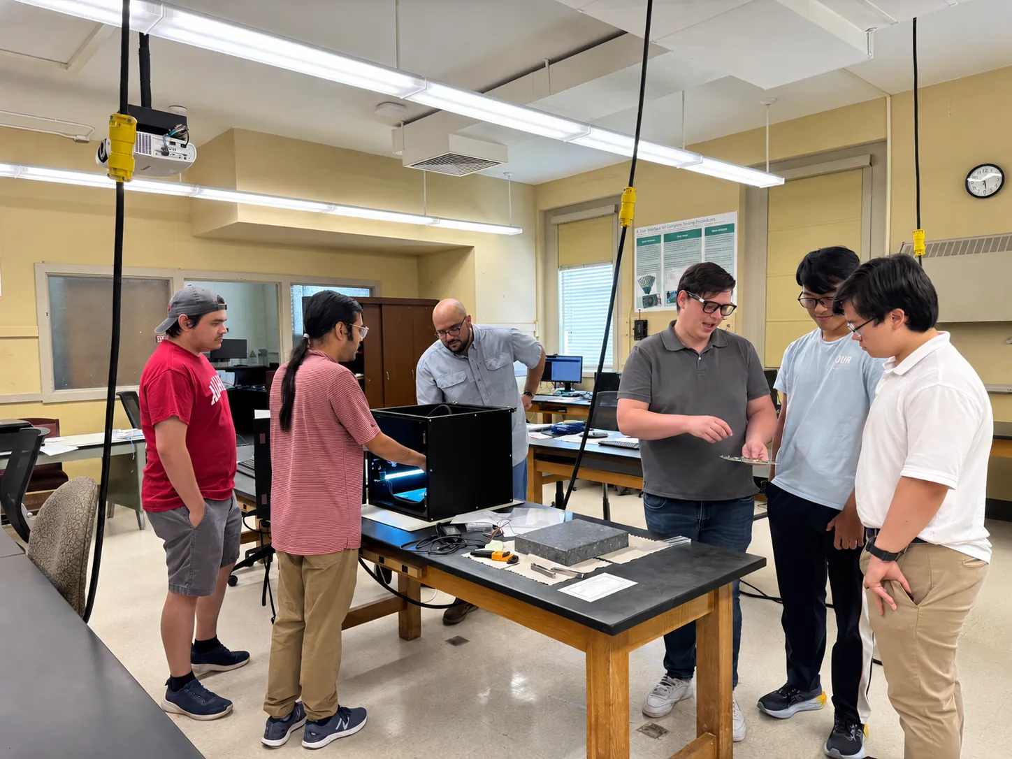 Six members of the group stand around a bench in the HGCAL lab; one holds up a hexaboard while the others look on. A black enclosure sits on the bench.