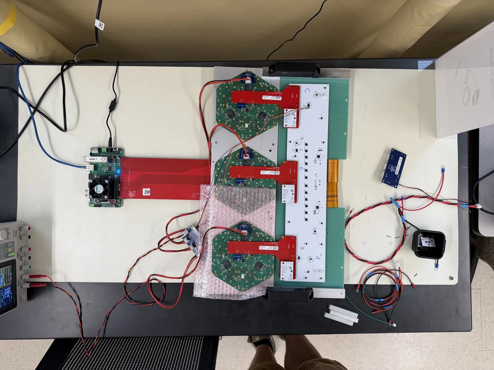 Top-down view of a test stand on a lab table: three green hexaboards, each with a red adapter board, connected to a long white board and to a small controller board at left, with a bench power supply at the edge of the table.